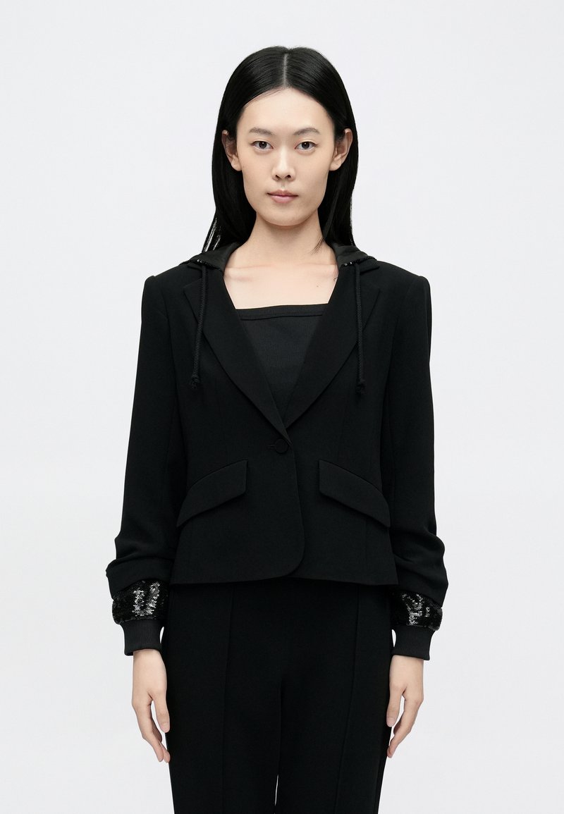 Mustast lõigatud bleiser kapuutsiga, esitaskud ja mustade litrite tekstuuriga mansetid. Sile kangas, istuv lõige.