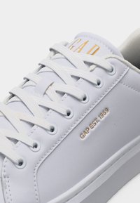 Baskets en cuir blanc avec lacets blancs, inscription dorée "GAP EST. 1969" sur le côté, et texte partiel "GAP" en or sur la languette.