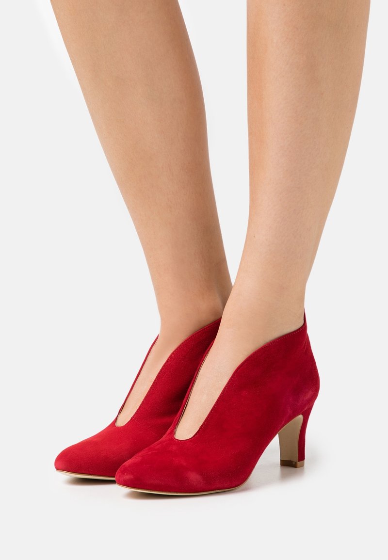 Paire de chaussures en daim rouge à talon moyen portées sur jambes nues, avec une coupe en V profonde et un talon beige empilé.