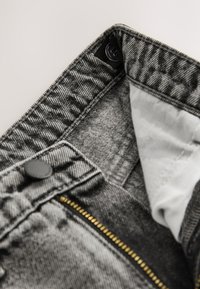 Grijze denim met een gestructureerde afwerking, voorzien van een messing rits en zwarte knoopsluiting. De voering aan de binnenkant is van lichtgekleurd materiaal voor contrast.