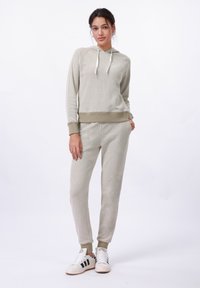 Giovane donna in piedi che indossa un hoodie beige e pantaloni sportivi abbinati con sneakers bianche, su uno sfondo bianco semplice.