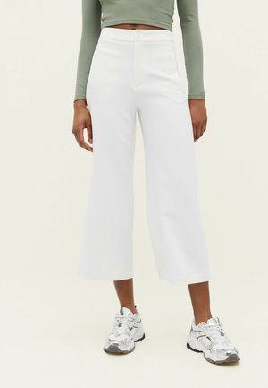 Person trägt weiße, weite, knöchellange Hosen, silberne Sportschuhe und ein grünes, langärmliges, kurzes Oberteil und steht vor einem hellen Hintergrund.