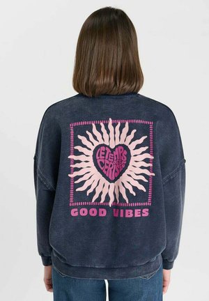 Persoon met schouderlang bruin haar die een donker sweatshirt draagt met een roze zon- en hartontwerp en de teksten "LETEMPS DES CERISES" en "GOOD VIBES".