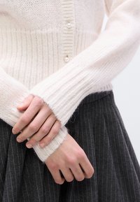 Ροζ ανοιχτή πλεκτή ζακέτα με κουμπιά από μαργαριτάρια, με ριμπ μανσέτες. Χέρια σταυρωμένα μπροστά, αναδεικνύοντας την απαλή υφή σε σύγκριση με φούστα με σκούρες ρίγες.