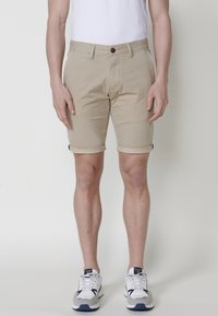 Beige cotton shorts with rolled cuffs, front button closure, and side pockets. Worn with white t-shirt and sporty sneakers.