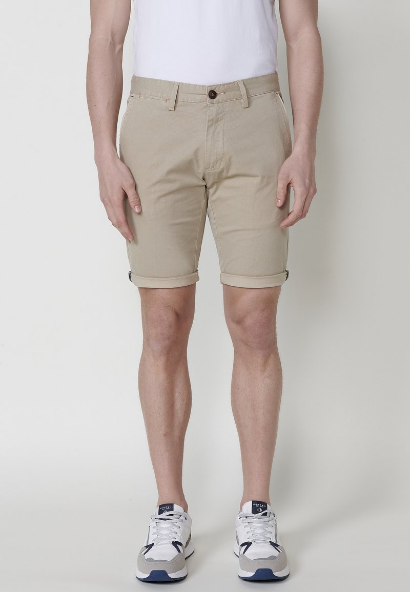 Beige cotton shorts with rolled cuffs, front button closure, and side pockets. Worn with white t-shirt and sporty sneakers.