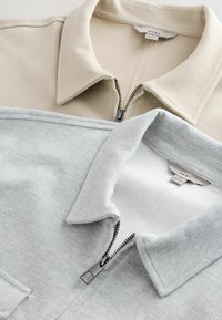 Dos chaquetas con cremallera: una beige con una tela texturizada y una gris claro en un material liso. Ambas tienen cuello y cremalleras visibles.