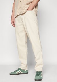 Pantalones de algodón beige con un corte relajado, que cuentan con bolsillos laterales y una textura suave. Combinados con zapatillas de gamuza verde y una camisa de estampado.
