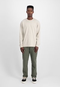 Beige lange mouw shirt met geribde manchetten, gecombineerd met olijfgroene broek met verticale logoprint, gedragen met zwart en crème sneakers.