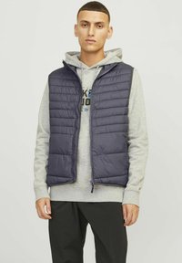 Marineblauwe puffer-vest met rits, voorzien van gequilt detail en een capuchon, gedragen over een grijze hoodie, gecombineerd met zwarte broek.
