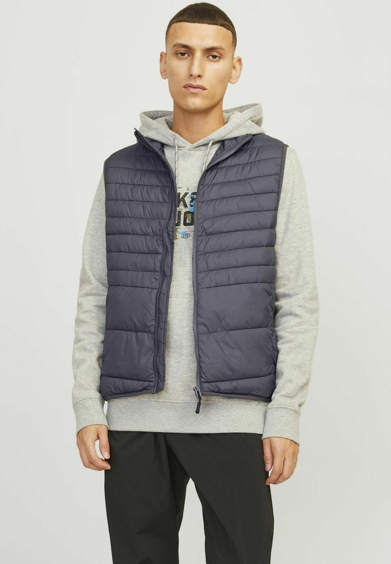 Marineblauwe puffer-vest met rits, voorzien van gequilt detail en een capuchon, gedragen over een grijze hoodie, gecombineerd met zwarte broek.