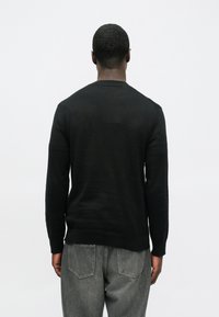 Homem de costas, vestindo uma camisa preta de manga longa e jeans cinzentos, contra um fundo branco liso.