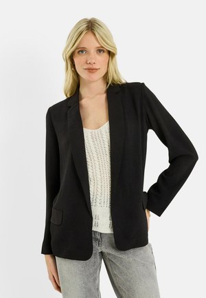 Femme blonde portant un blazer noir par-dessus un top en maille blanc et un jean gris clair, debout avec une main sur la hanche.