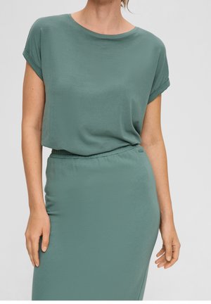 Femme portant une robe ajustée vert sauge à manches courtes avec taille élastique, debout devant un fond uni gris clair.
