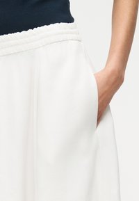 Personne portant un pantalon blanc à taille élastique avec la main dans la poche latérale et un haut foncé visible à la taille.