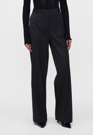 Femme portant un cardigan noir transparent boutonné, un pantalon large à fines rayures, des chaussures noires pointues et des lunettes de soleil rondes, debout devant un fond blanc.