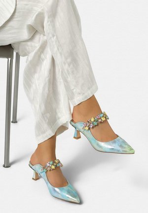 Legs in white pants wearing shiny blue-green pointed-toe kitten heels with multicolored gem embellishments across the top straps.