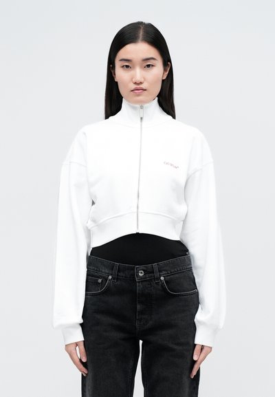 Sweatshirt bianco corto con colletto alto e chiusura con zip, dotato di maniche lunghe e larghe e ricami sottili in rosso. Abbinato a jeans neri.