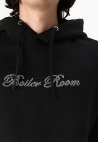 Black hoodie made of soft fabric featuring a drawstring hood. It has "Boiler Room" embellished with rhinestones across the front.
