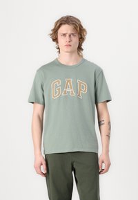 Žalias medvilnės marškinėlis su dideliu smėlio spalvos "GAP" logotipu ir tekstu "Athletic" po juo. Trumpomis rankovėmis, apvalia iškirpte, laisvas siluetas.