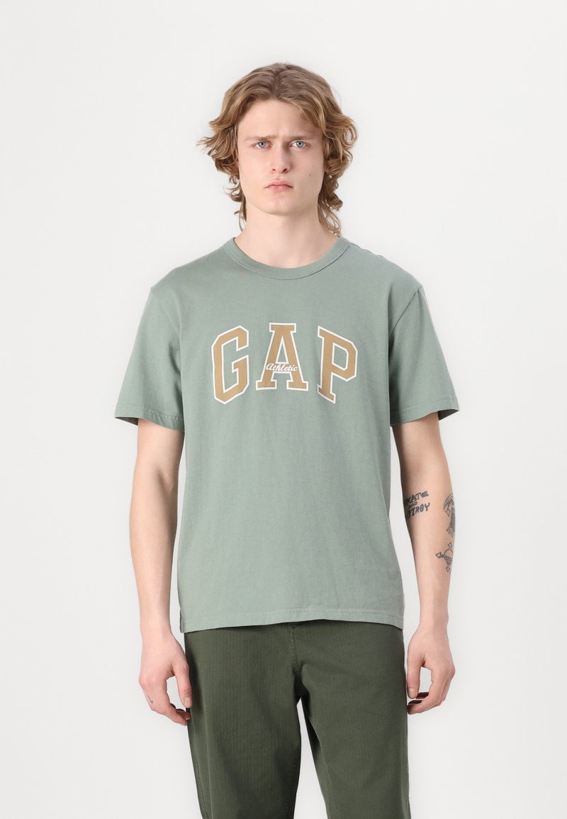 Žalias medvilnės marškinėlis su dideliu smėlio spalvos "GAP" logotipu ir tekstu "Athletic" po juo. Trumpomis rankovėmis, apvalia iškirpte, laisvas siluetas.