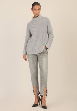 Grauer Rippstrickpullover mit hohem Kragen, kombiniert mit hellgrauen Jeans mit ausgefransten Säumen und Perlenakzenten. Schwarze High Heels vollenden das Outfit.