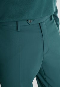 Tirkizne hlače izrađene od glatkog materijala s krojem koji odgovara tijelu, s ravnim prednjim dijelom, petljama za remen i jednim dugmetom za zatvaranje.