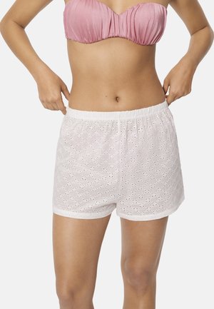 Mujer con un top sin tirantes rosa y pantalones cortos blancos con bordado floral, ajustándose la cintura a ambos lados.