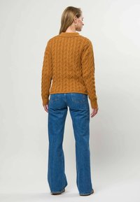 Femme de dos portant un pull marron en maille torsadée et un jean large bleu, debout devant un fond clair uni.