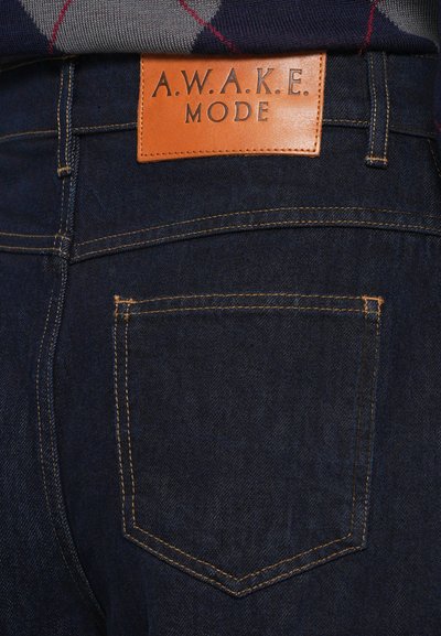 Tumedega asendiga teksapüksid, tume sinine värv, millel on oranž õmblus ja helepruun nahklabel, millel on reljeefne kirjutis "A.W.A.K.E. MODE."