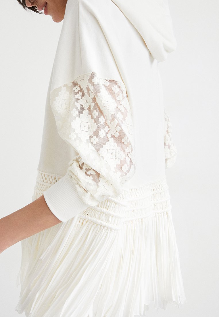 Felpa bianca con maniche bordate in pizzo e orlo frangiato. Presenta un tessuto testurizzato e intricate fantasie in pizzo sulle maniche.