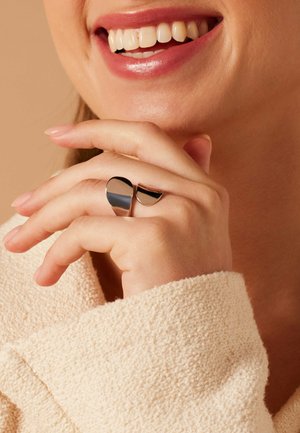 Lächelnde Frau mit einem strukturierten beigen Pullover zeigt einen silbernen abstrakten offenen Ring an ihrer rechten Hand nahe dem Gesicht.