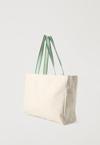 Μπεζ ορθογώνια τσάντα ώμου με λουράκια κρέμας με πράσινες άκρες, που στέκεται ενάντια σε ένα απλό λευκό φόντο.