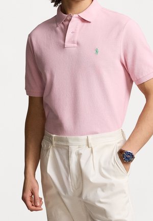 Homme portant un polo à manches courtes rose clair avec un logo vert, un pantalon plissé crème et une montre au cadran bleu avec un bracelet en cuir marron.