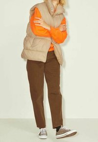 Beige vadderad väst över en orange sweatshirt, i kombination med bruna kordflöjlsbyxor och grå sneakers med vita sålar. Enkelt mönster och varma färger.