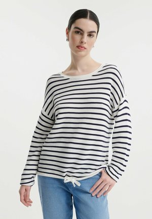 Langärmliges gestreiftes Oberteil in Creme und Marineblau, mit runden Ausschnitt, lockerer Passform und Kordelzugdetails am Saum. Kombiniert mit Jeans.