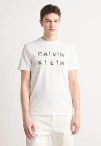 White cotton t-shirt featuring bold black "CALVIN KLEIN" text in a modern font. Classic crew neckline and short sleeves. Smooth texture.