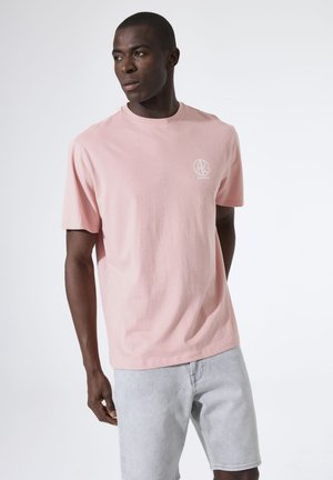 Mann trägt ein hellrosa kurzärmeliges T-Shirt mit kleinem weißem Logo auf der Brust und hellgraue Denim-Shorts, steht und schaut nach links.