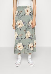 Bloemenrokken in gedempte groene tint met beige en marineblauwe bloemmotieven. Elastische tailleband, enkellang, gecombineerd met witte sneakers.