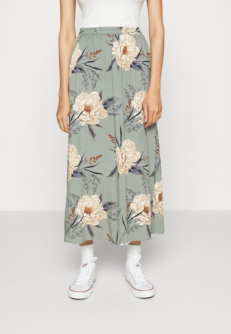 Bloemenrokken in gedempte groene tint met beige en marineblauwe bloemmotieven. Elastische tailleband, enkellang, gecombineerd met witte sneakers.