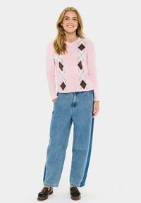 Rosa Strickjacke mit Knopfverschluss und Argyle-Muster in Braun, Blau und Weiß. Kombiniert mit übergroßen blauen Jeans und braunen Schuhen.
