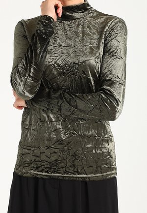 Person iført en langærmet, højhalset, skinnende grøn crushed velvet-top med sort underdel, poserer med én hånd tæt ved ansigtet.