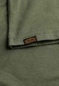 Oliven grønt stof med et sort og orange Superdry Outfitters mærke syet nær den foldede kant.