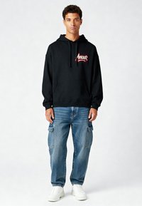 Felpa nera con logo "Amour" rosso, tasca a marsupio, abbinata a jeans cargo azzurri chiari e sneakers bianche. Tessuto morbido, vestibilità rilassata.