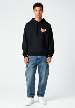 Zwarte hoodie met "Amour" logo in het rood, kangoeroezak, gedragen met lichtblauwe cargobroek en witte sneakers. Zachte stof, ontspannen pasvorm.