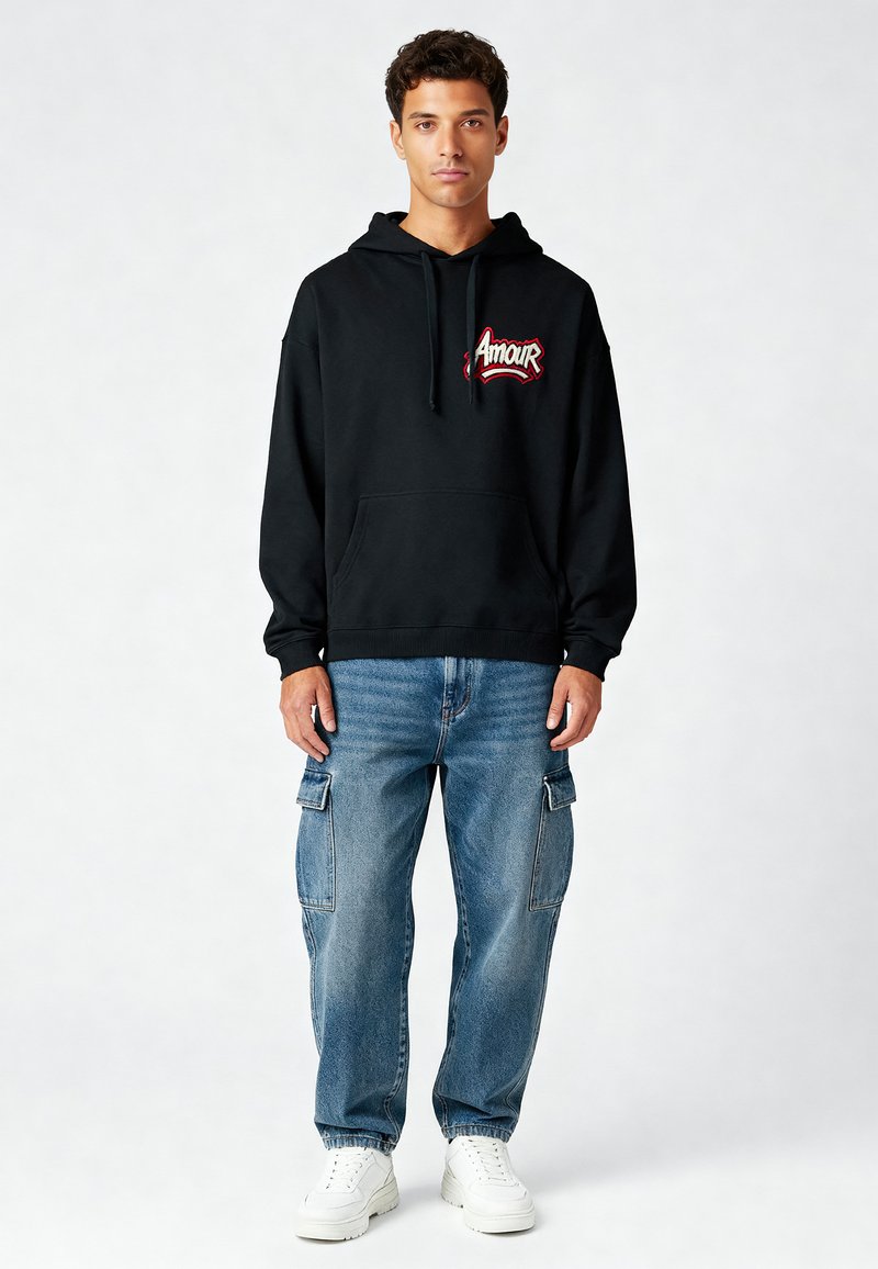 Sweat à capuche noir avec le logo "Amour" en rouge, poche kangourou, associé à un jean cargo bleu clair et des baskets blanches. Tissu doux, coupe décontractée.