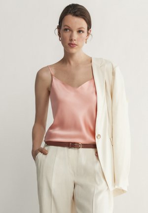 Camisole en satin rose avec de fines bretelles, associé à un blazer blanc et un pantalon beige clair, accentué par une ceinture marron. Design minimaliste.