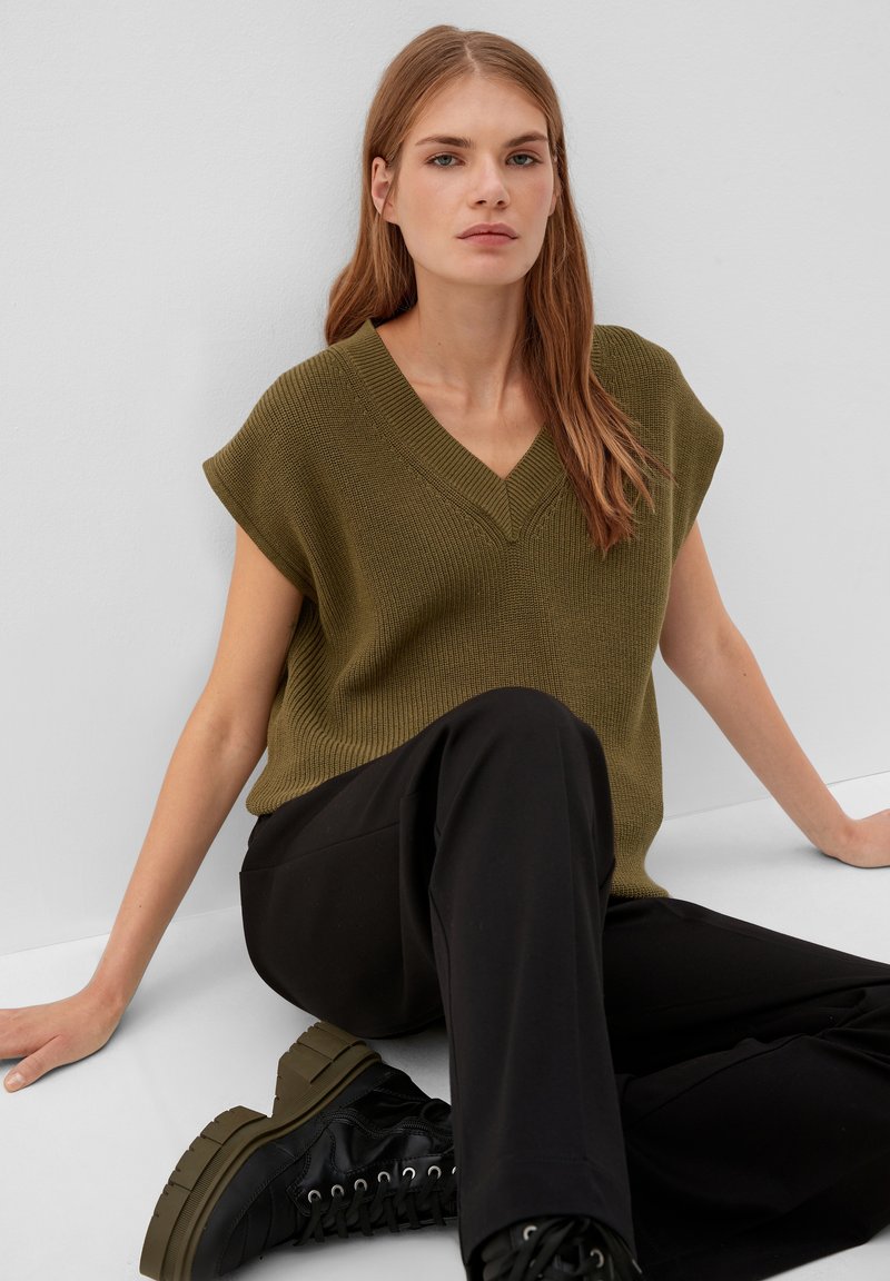 Olivgrünes, gestricktes ärmelloses Oberteil mit V-Ausschnitt, kombiniert mit schwarzen, weit geschnittenen Hosen und schwarzen, klobig-sohligen Stiefeln. Einfaches Design.