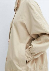 Beige jacket with a relaxed fit, featuring a high collar, elastic cuffs, and side pockets. Smooth fabric with a matte finish.
