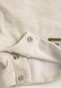 Tissu en coton beige clair avec une finition texturée, comportant des fermetures pression argentées et une étiquette marron attachée près de l'ourlet.